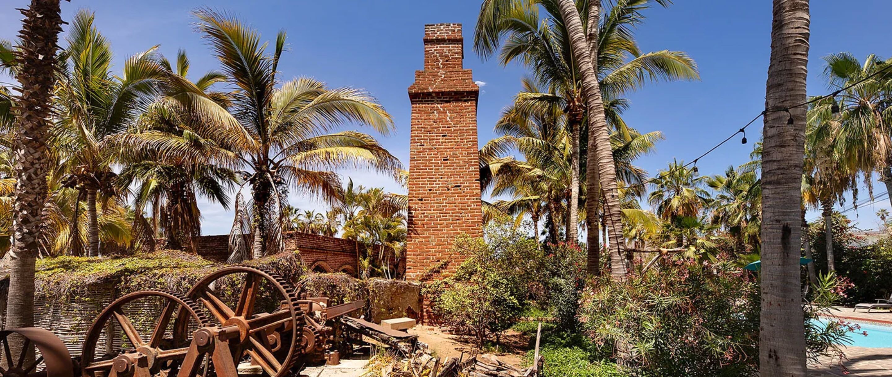 An old brick structure with a chimney, surrounded by palm trees and a large water wheel.