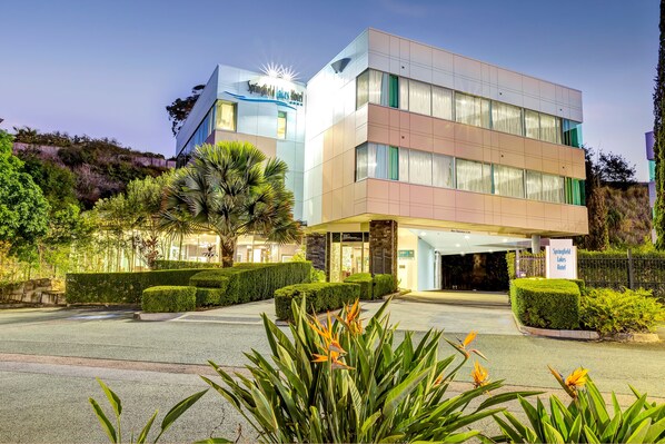 A modern hotel with a two-story structure, a well-maintained garden, and a clear sky.