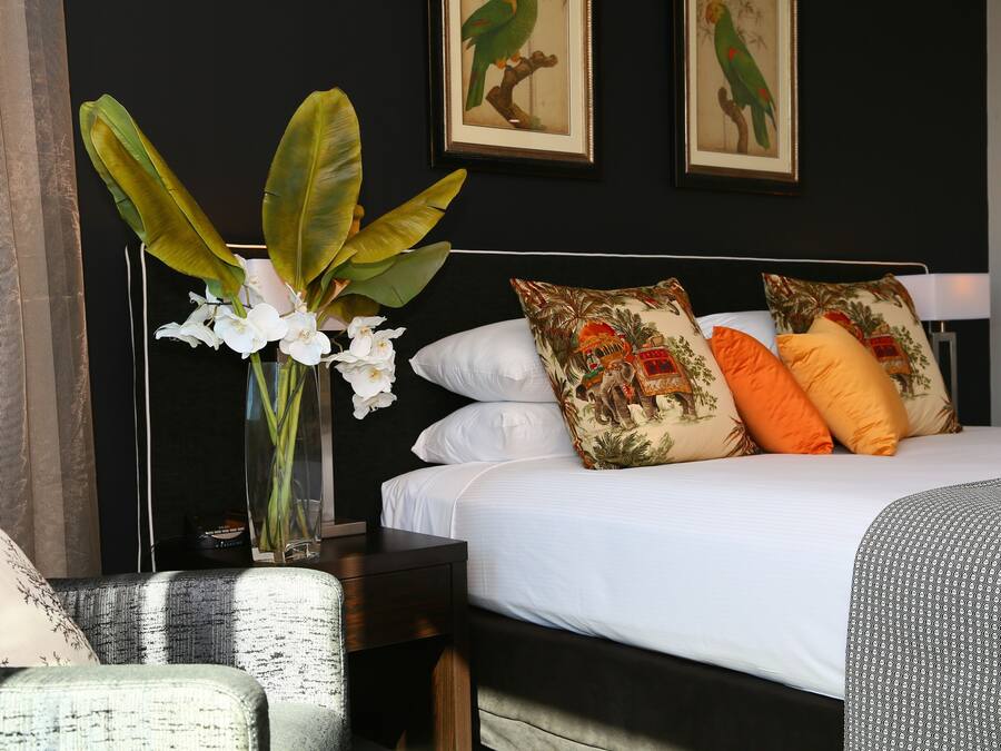 Een netjes opgemaakt bed met witte lakens en gestreepte kussens, een vaas met witte bloemen, en een lijst met een foto van een papegaai aan de muur.