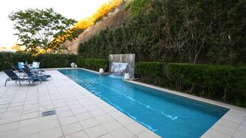 A swimming pool with a waterfall feature, surrounded by neatly trimmed hedges and a tiled deck with lounge chairs.