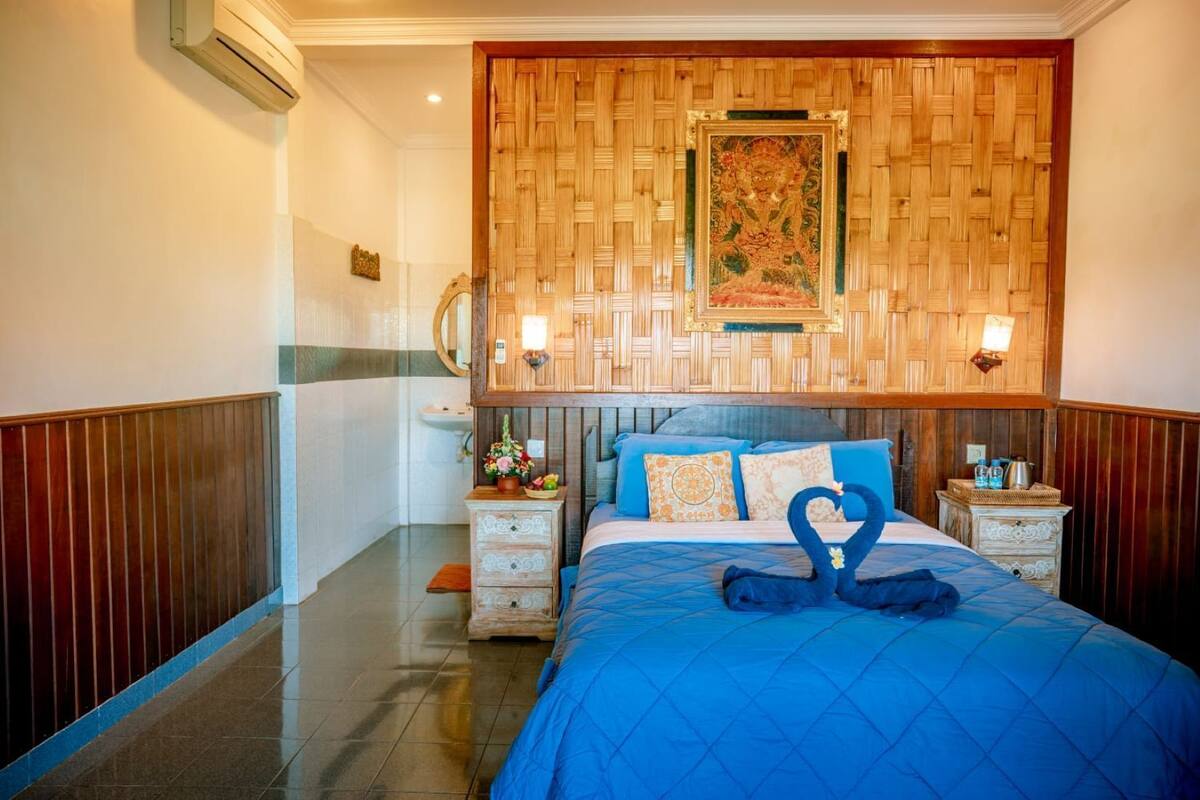 A hotel room with a large bed, two bedside tables, a wooden headboard, a decorative tapestry, and a wall-mounted air conditioning unit.