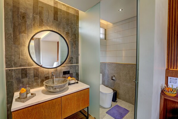Un baño moderno con pared de baldosas de piedra, un espejo redondo, tocador de madera, lavabo de piedra y un inodoro.