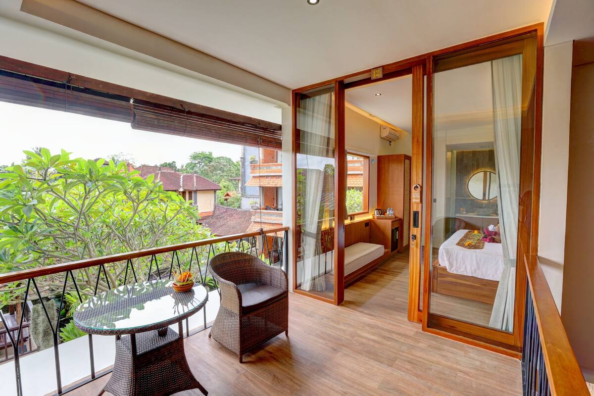 A balcony with a wicker chair and table, a sliding glass door leading to a bedroom with a bed and a small table.