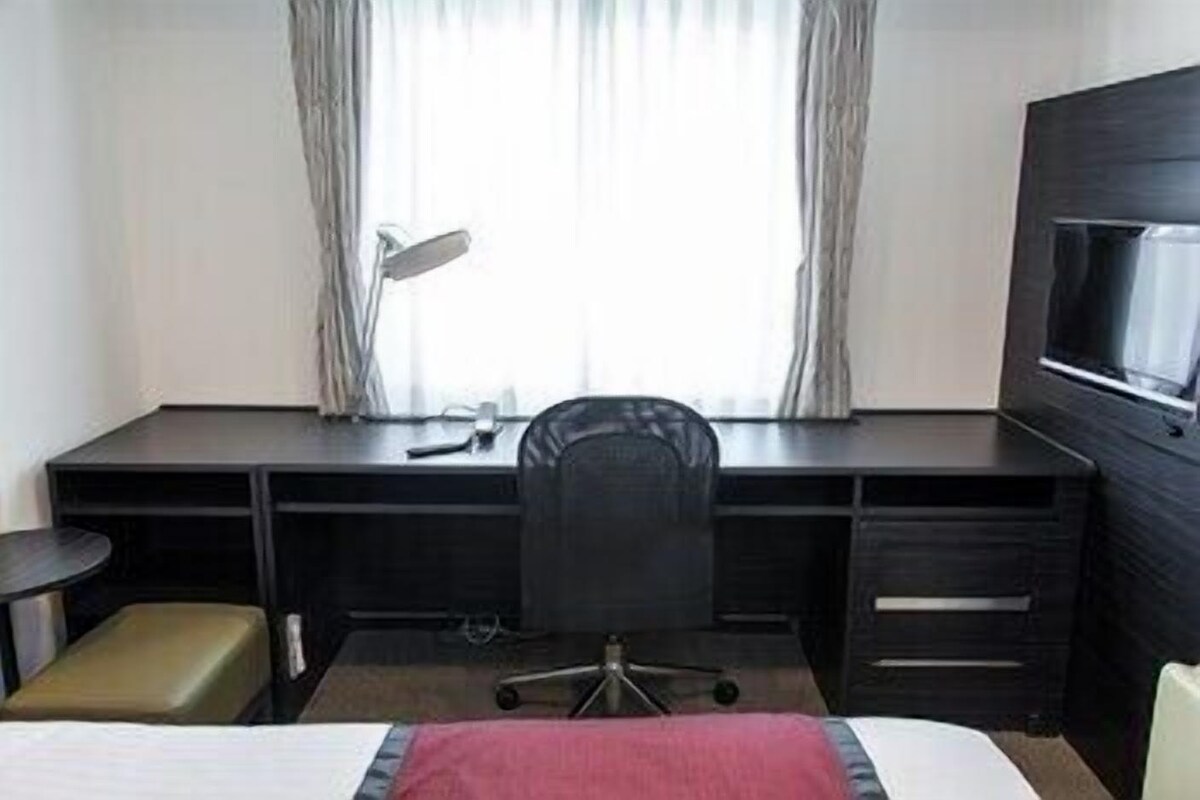 A hotel room with a bed, a desk with a chair, a TV, and a window with curtains.