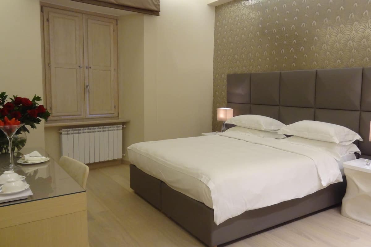 Habitación de hotel con cama, mesitas de noche, un radiador, una ventana, una mesa con un florero y una silla.