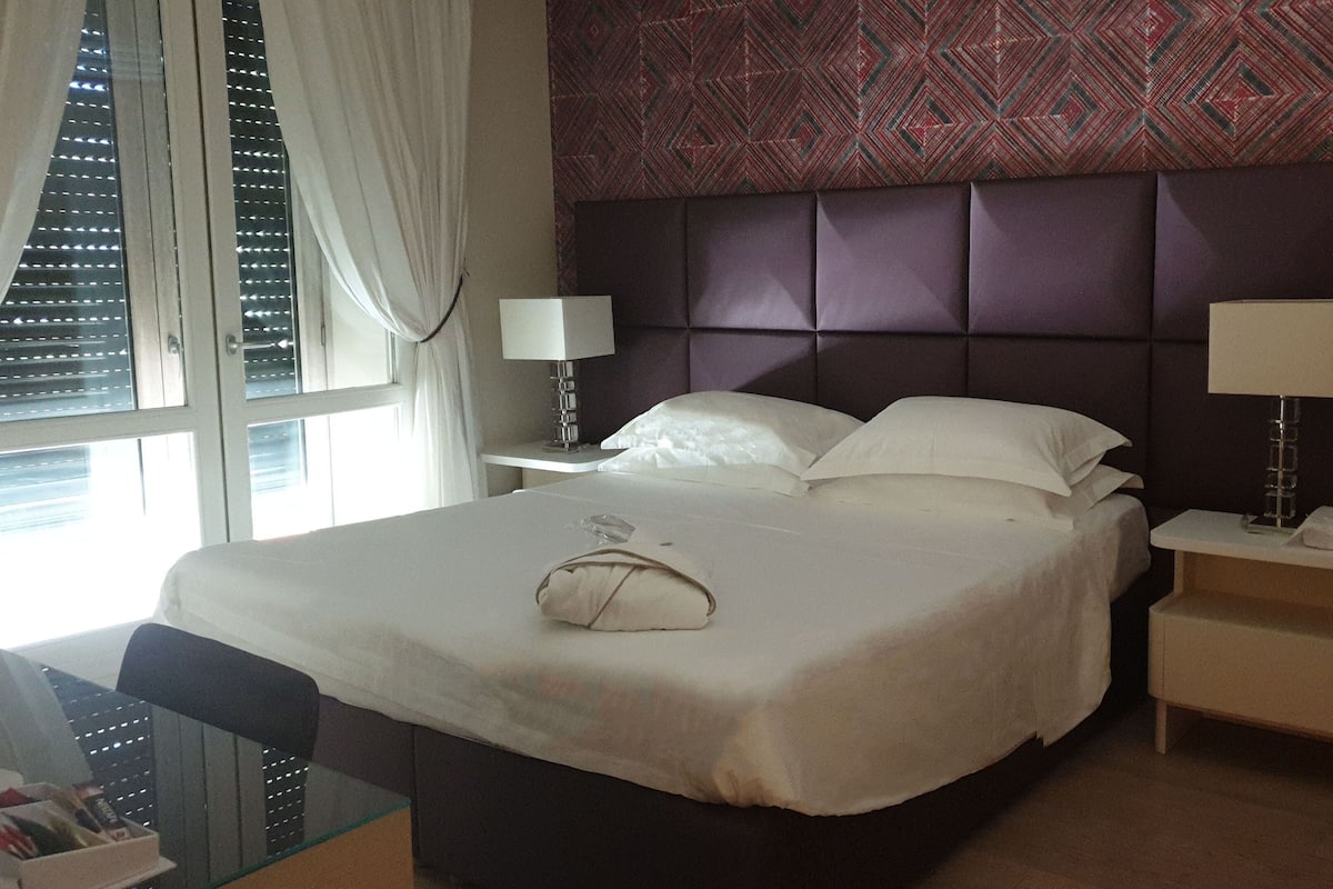 Una habitación de hotel con una cama grande, dos mesitas de noche con lámparas, un ventanal con persianas y una pared con diseño.