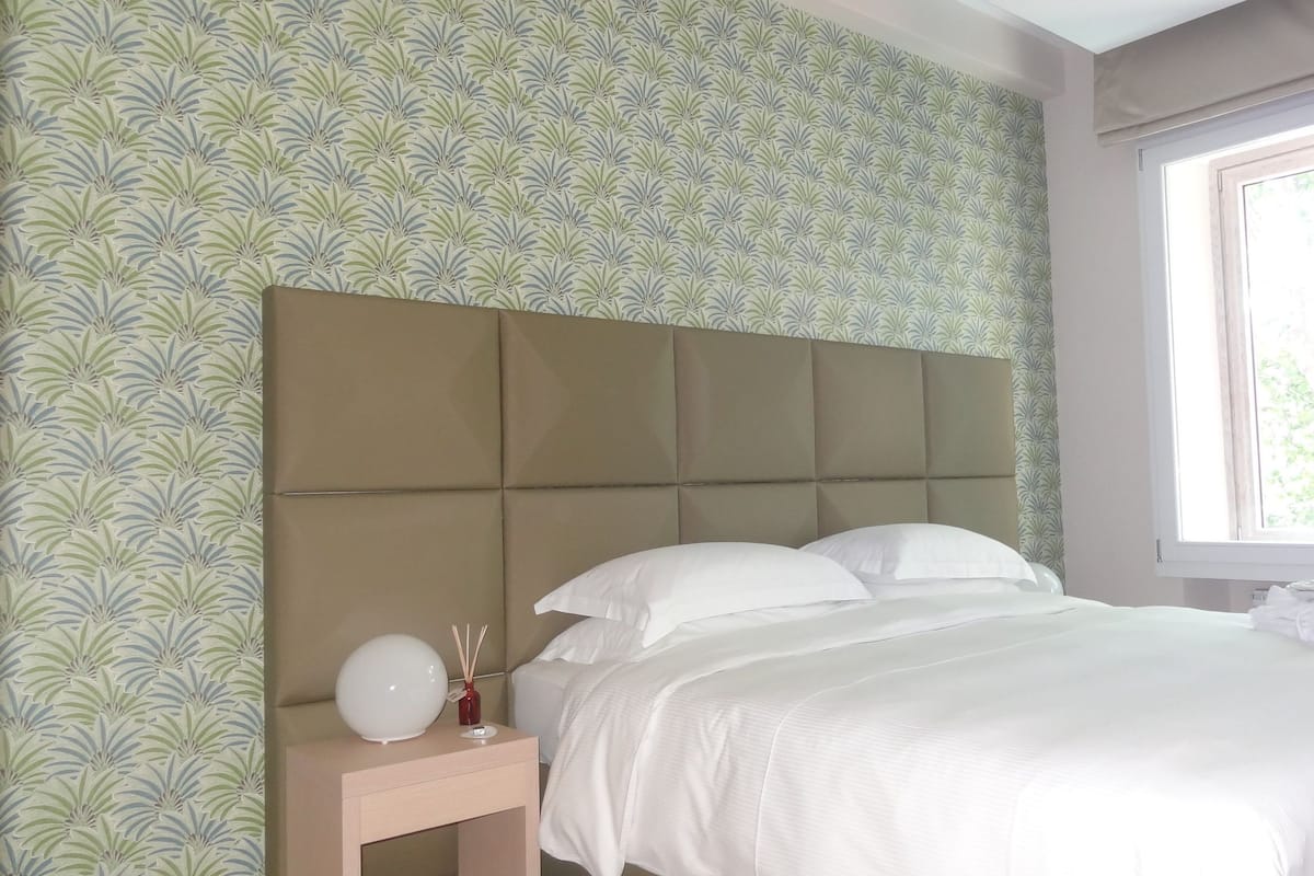 Un dormitorio con cabecera, una mesita de noche con lámpara y un florero, y una ventana con cortinas.