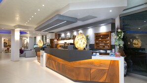 A modern reception desk with a wooden finish, a large decorative light fixture, and a view of the lobby area with seating and a bar. - Hotel Metropol (Chioggia)