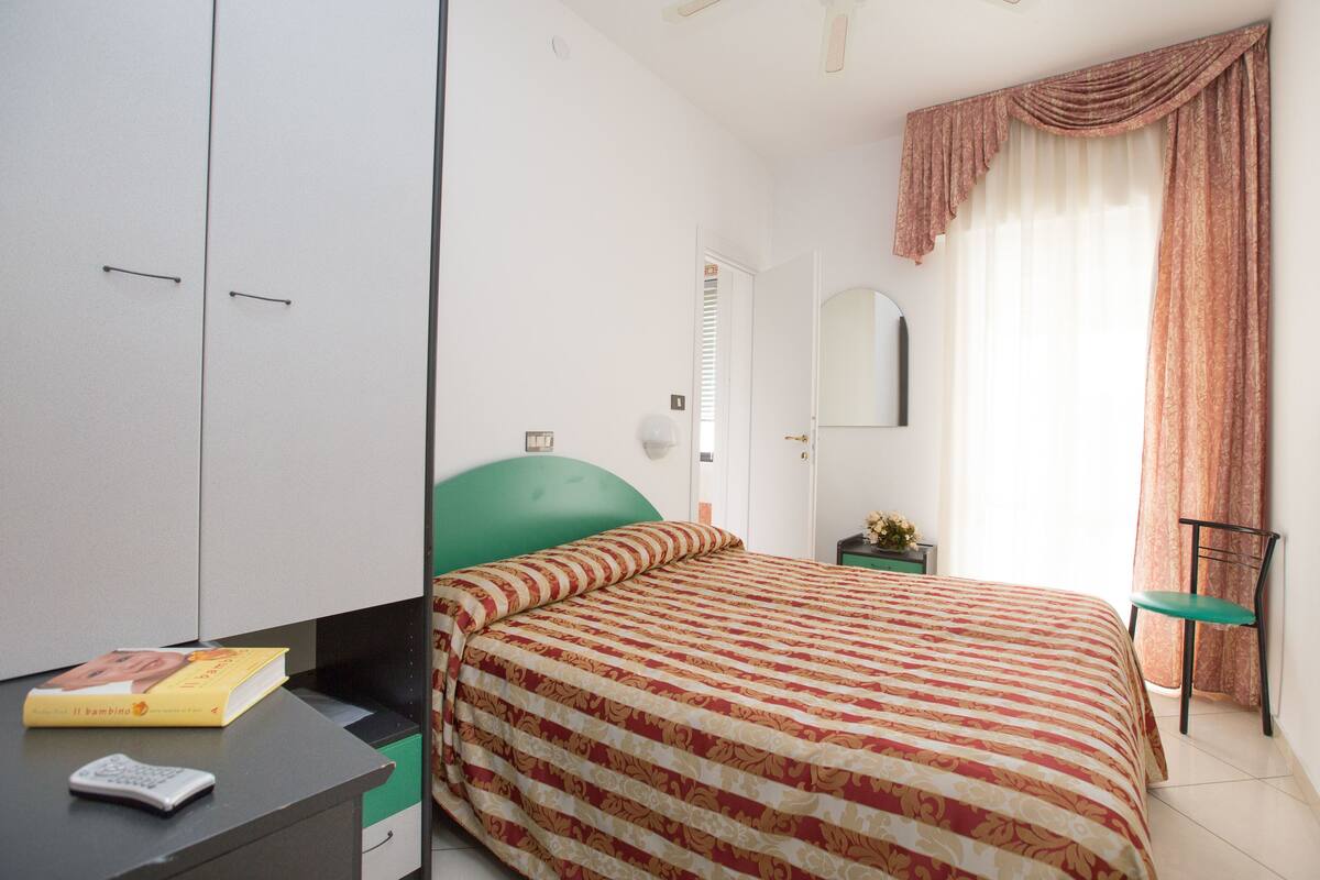 A bedroom with a bed, a desk with a book and remote, a green chair, and a window with curtains.