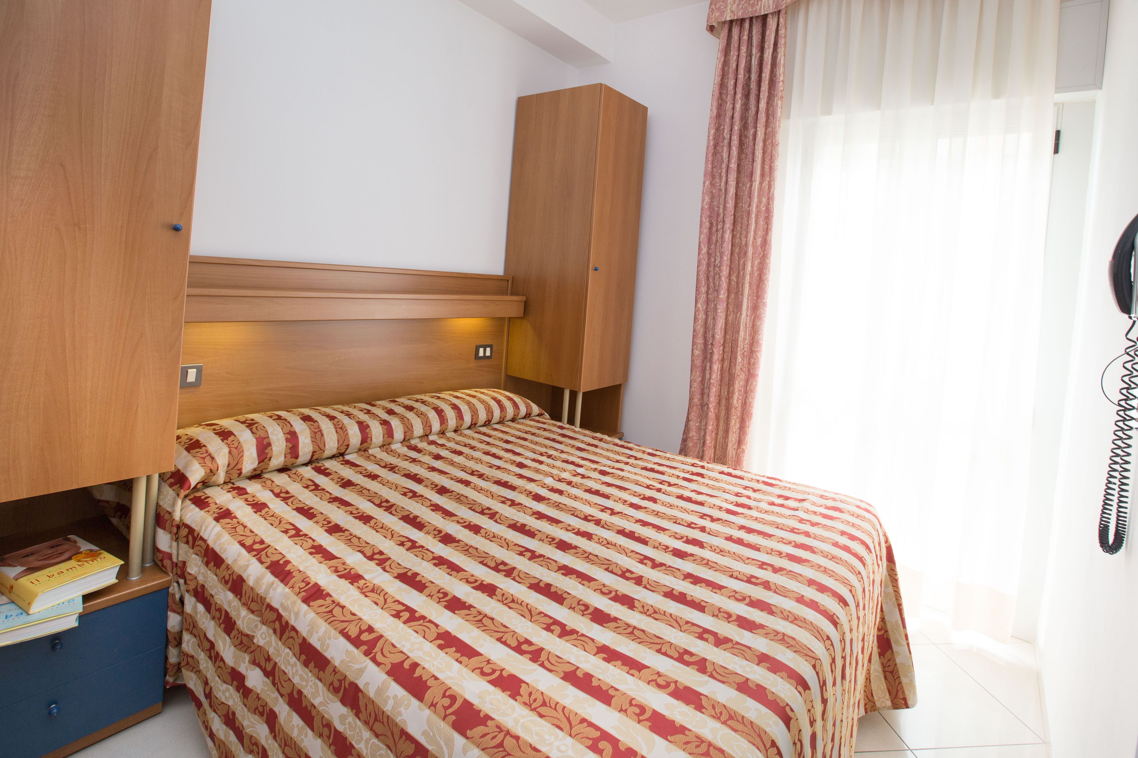 A bedroom with a bed, a wardrobe, a bedside table with books, and a telephone.