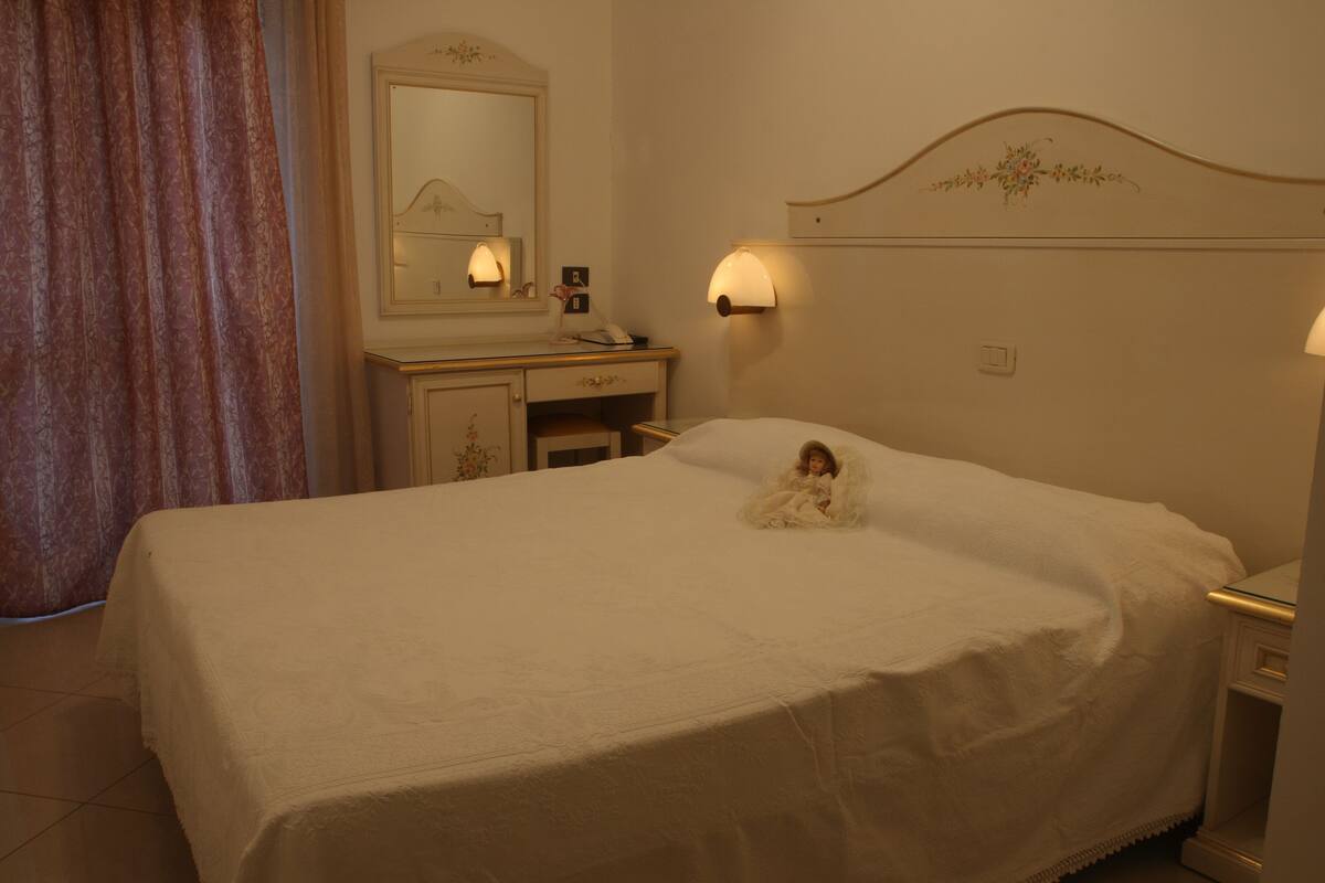 A neatly made bed with a small doll on it, a bedside table with a lamp, and a mirror above the dresser.