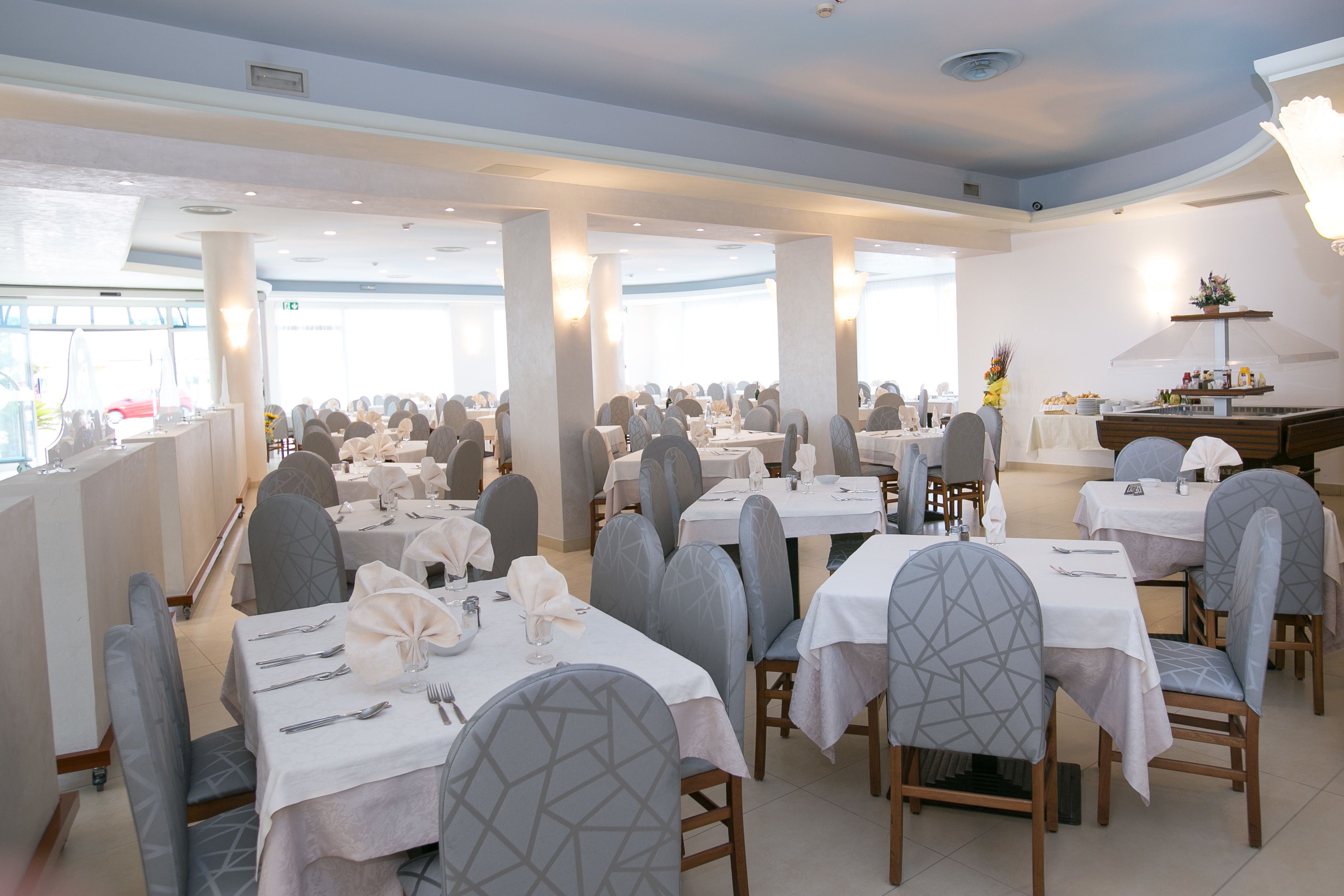 Un comedor amplio con varias mesas y sillas, ventanas grandes y una zona central tipo buffet.