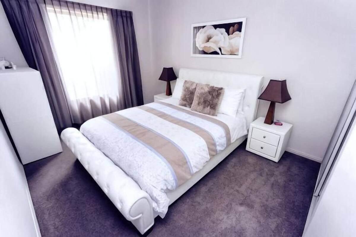 Un dormitorio ordenado con cama, mesitas de noche, una lámpara y un cuadro en la pared.
