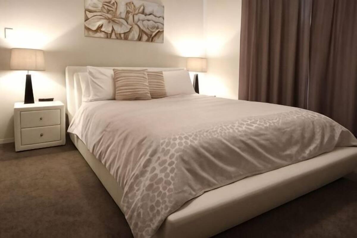 Una cama bien hecha con una colcha estampada, dos lámparas de noche y una mesita de noche con cajones.