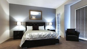 A bedroom with a large bed, two bedside tables, a chair, a mirror, and a window with blinds.