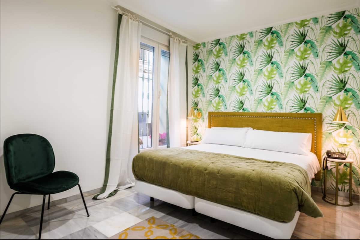 Un dormitorio con cama, una silla, una ventana con cortinas y un muro con diseño de hojas.