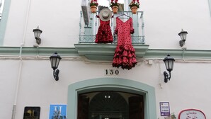 Un edificio con un balcón decorado con vestidos estampados en rojo y negro, el número '130' sobre la entrada y dos macetas a cada lado.