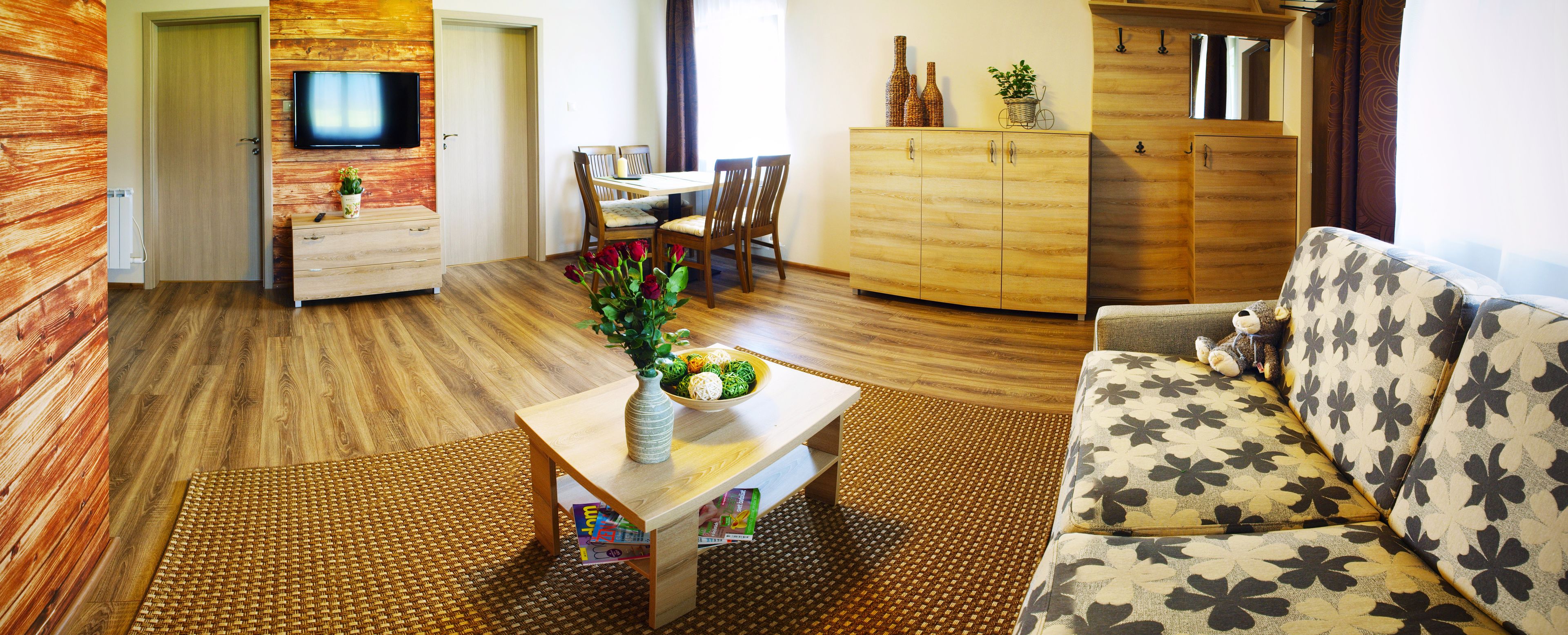 Een woonkamer met een bank, een bijzettafeltje met een schaal en bloemen, een eettafel met stoelen, een aan de muur bevestigde televisie en houten kasten.