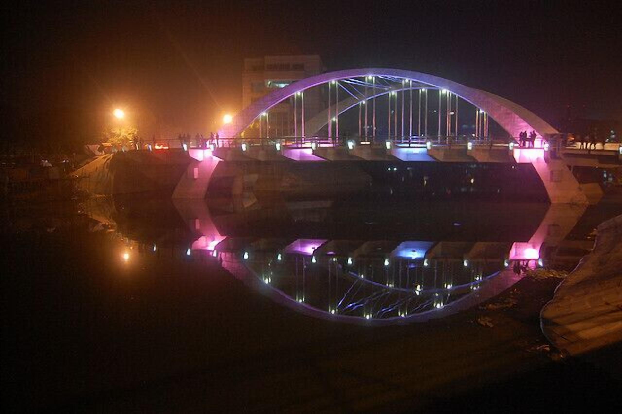 A modern bridge with a unique arch design, illuminated by colorful lights, spanning over a body of water.