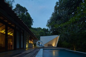 Un área de piscina moderna al aire libre con un elemento acuático geométrico, una terraza de madera y un edificio tradicional con puertas correderas.