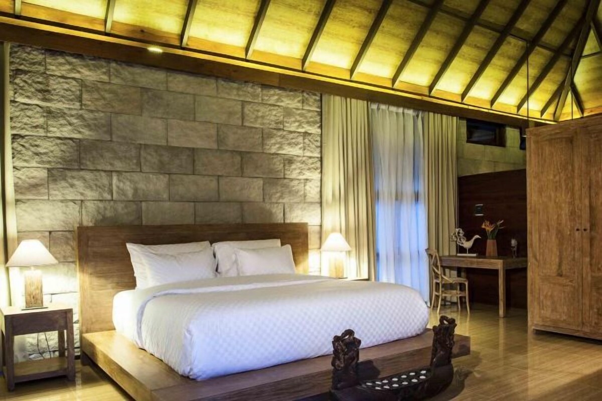 A bedroom with a large bed, a wooden headboard, a stone wall, a nightstand, a lamp, and a wooden cabinet.