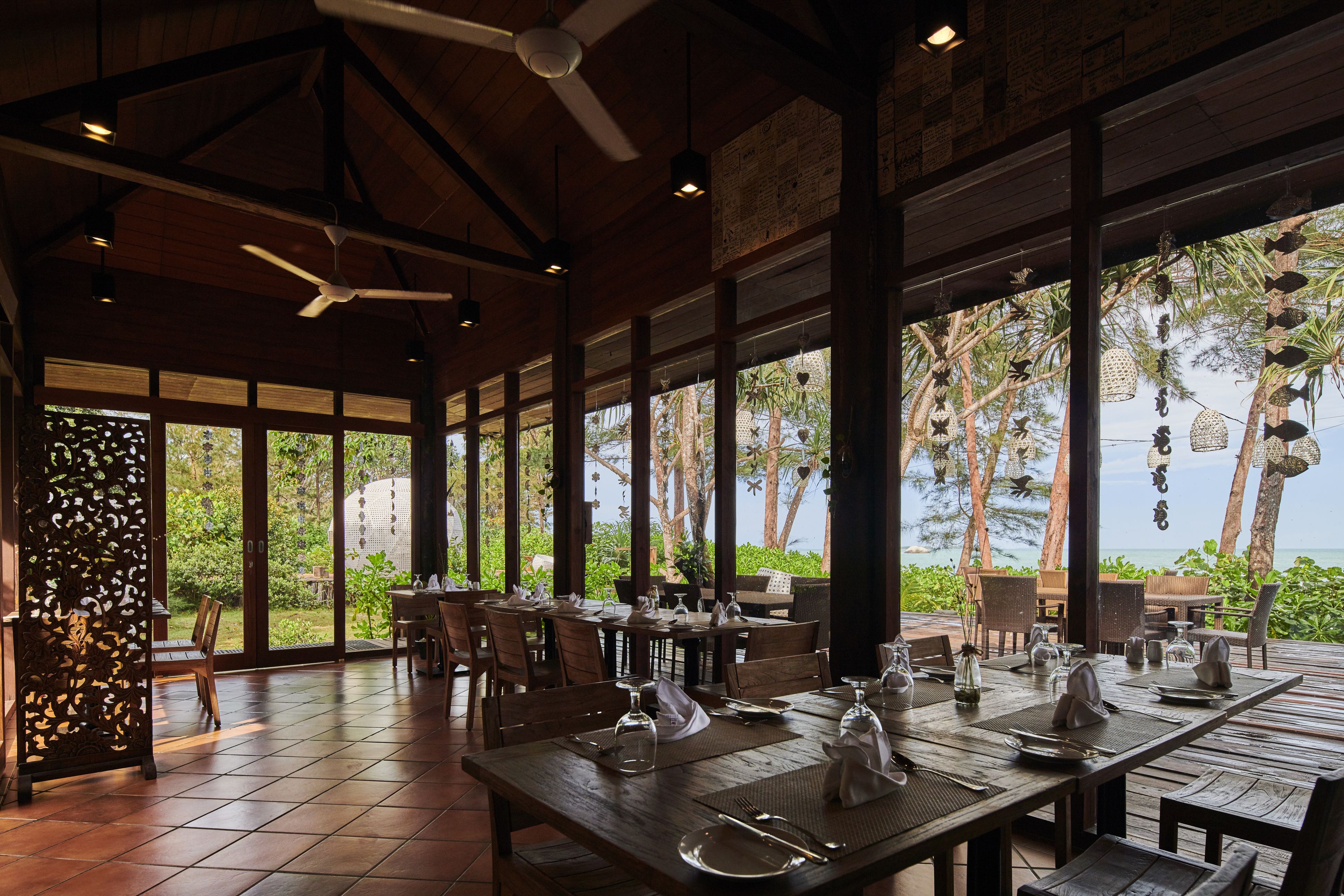 Un restaurant avec des tables et des chaises en bois, de grandes fenêtres et des ventilateurs de plafond.