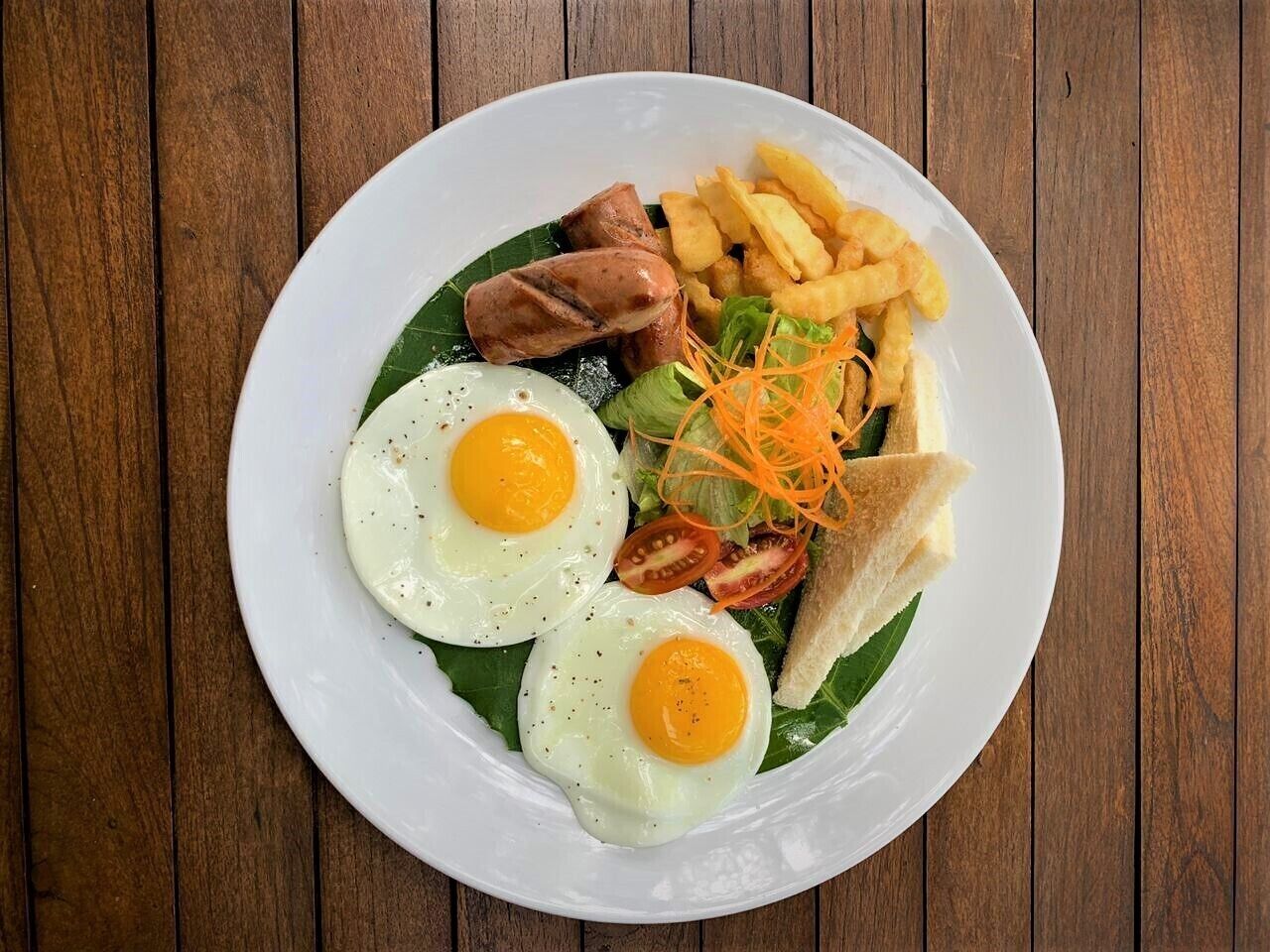 A plate of food with eggs, sausages, fries, and vegetables on a wooden table.
