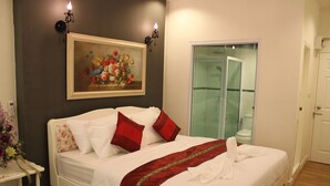 Un lit bien fait, avec du linge de lit blanc et des coussins rouges, un tableau de fleurs accroché au mur, et une salle de bain visible à travers une porte en verre.