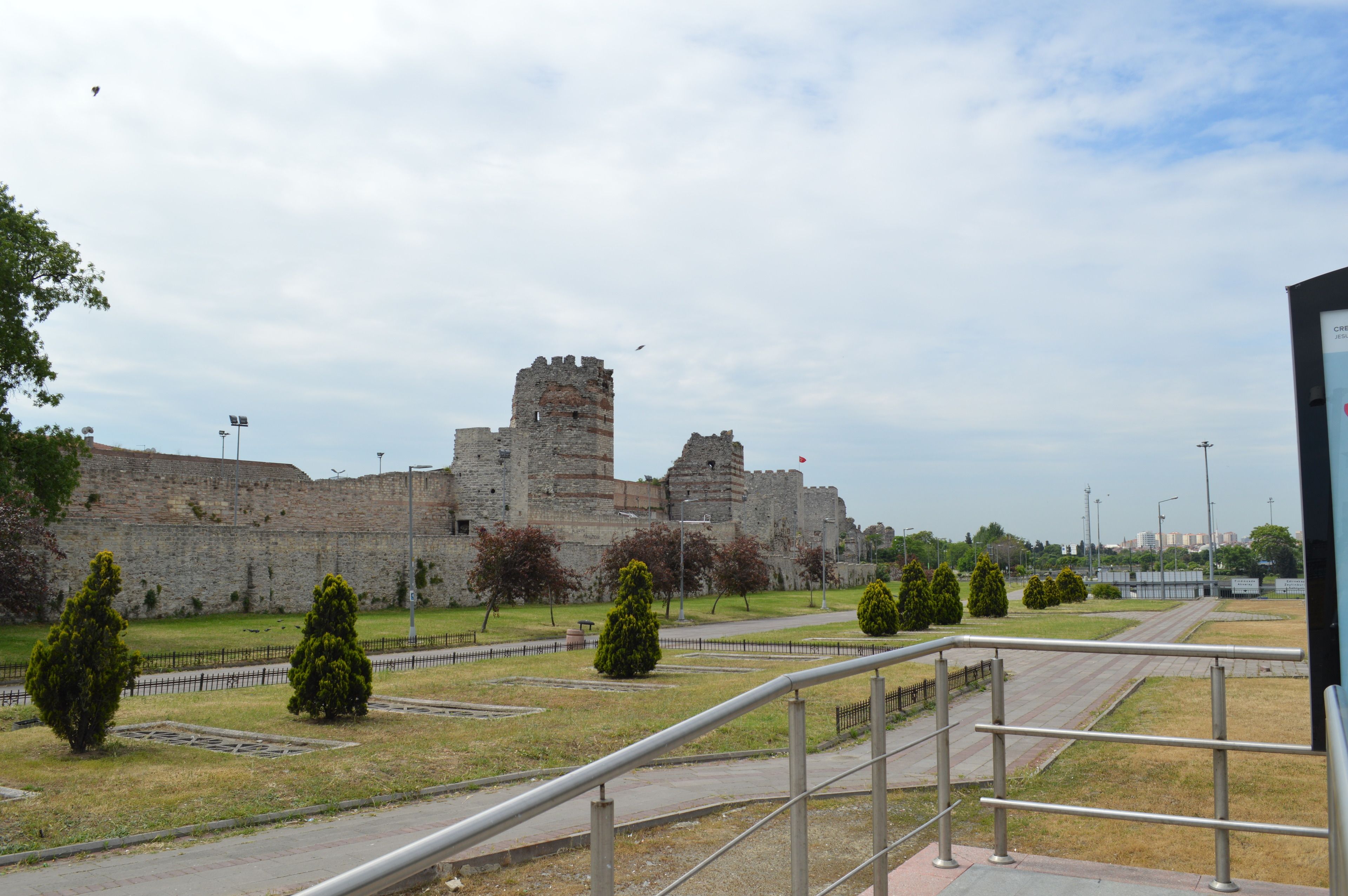 A historic stone castle with a central tower, surrounded by a paved area with pathways and trimmed hedges.