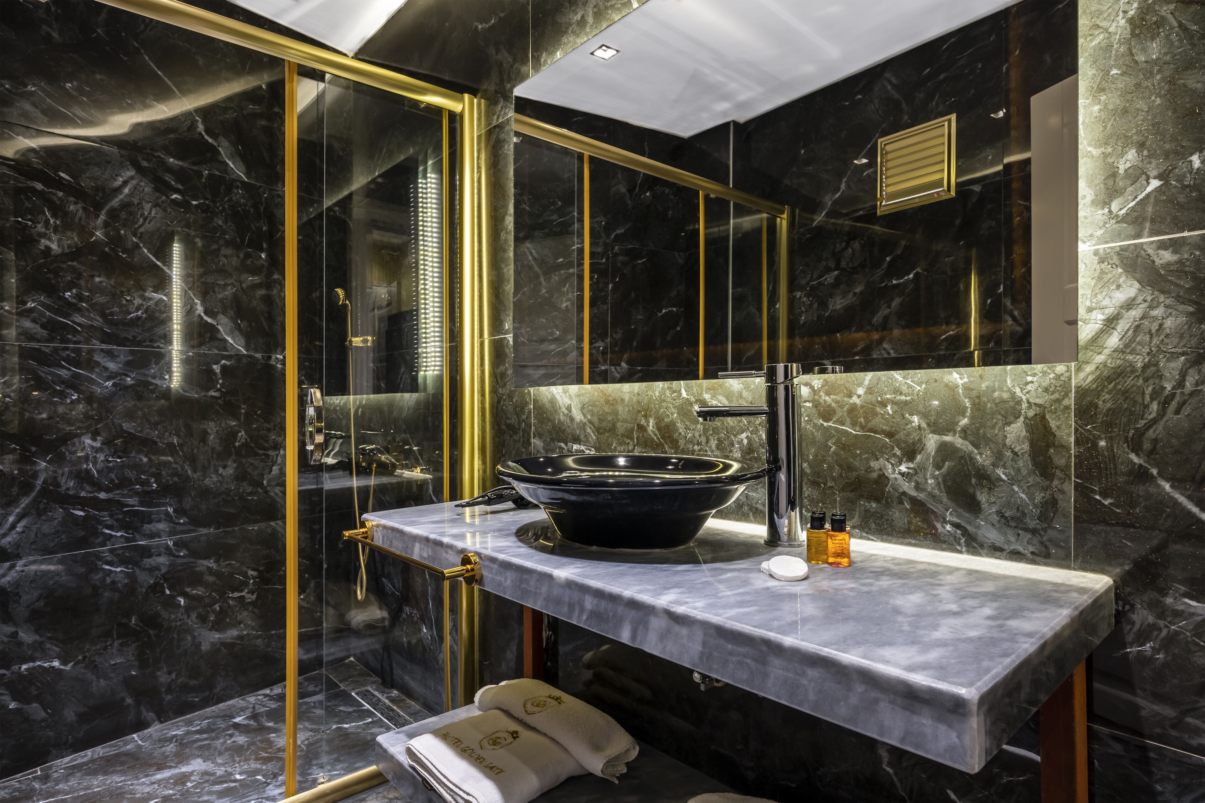 Un baño moderno con lavamanos de mármol negro, detalles dorados y ducha con cerramiento de vidrio.