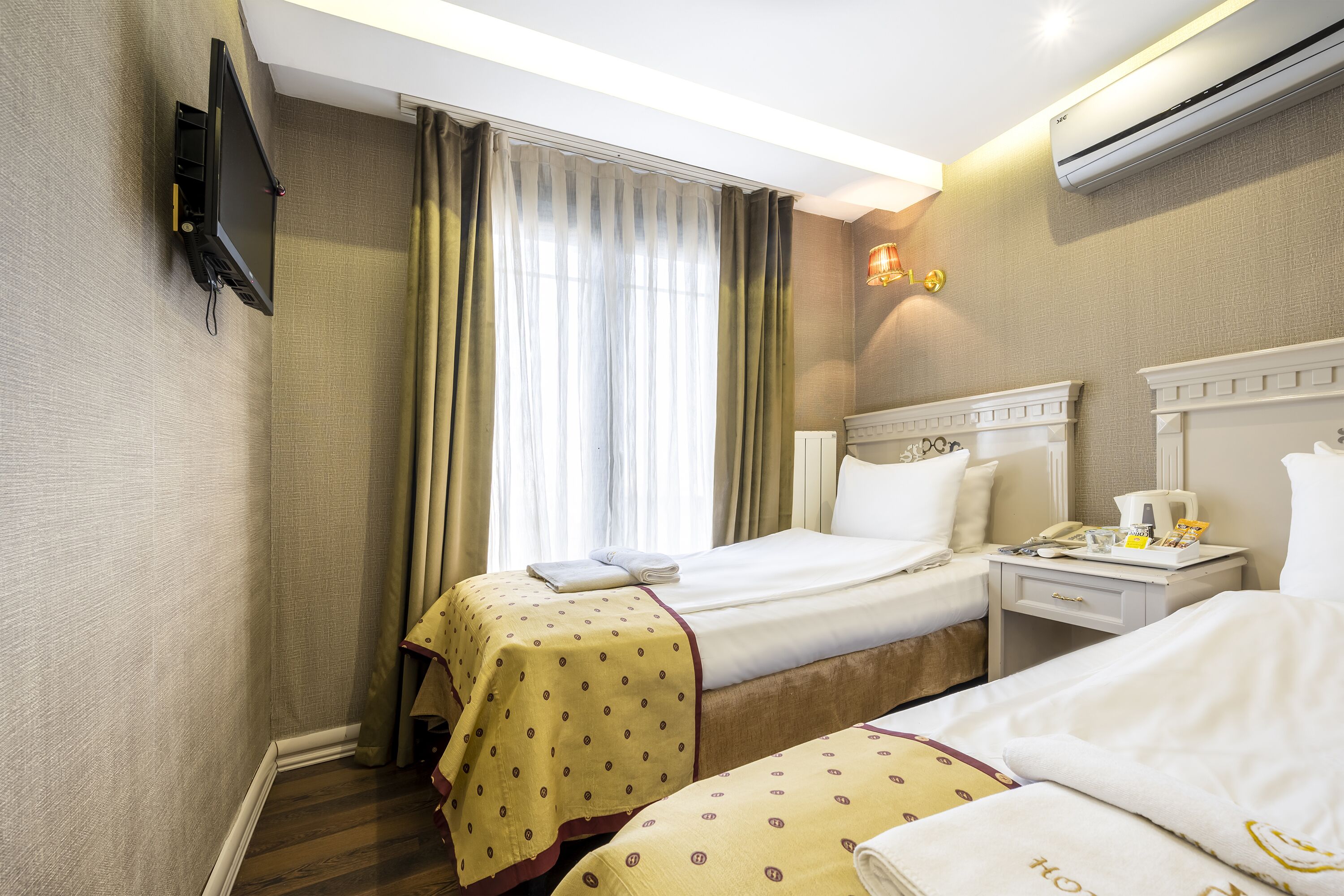 A hotel room with two beds, a TV mounted on the wall, a bedside table with a lamp, and an air conditioning unit.