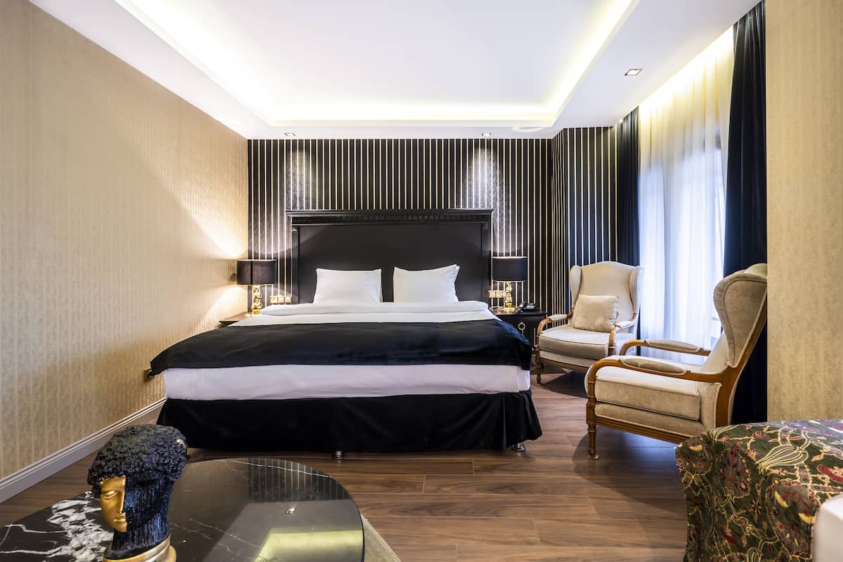 A hotel room with a large bed, a sitting area with a chair, a small table, and a decorative bust.