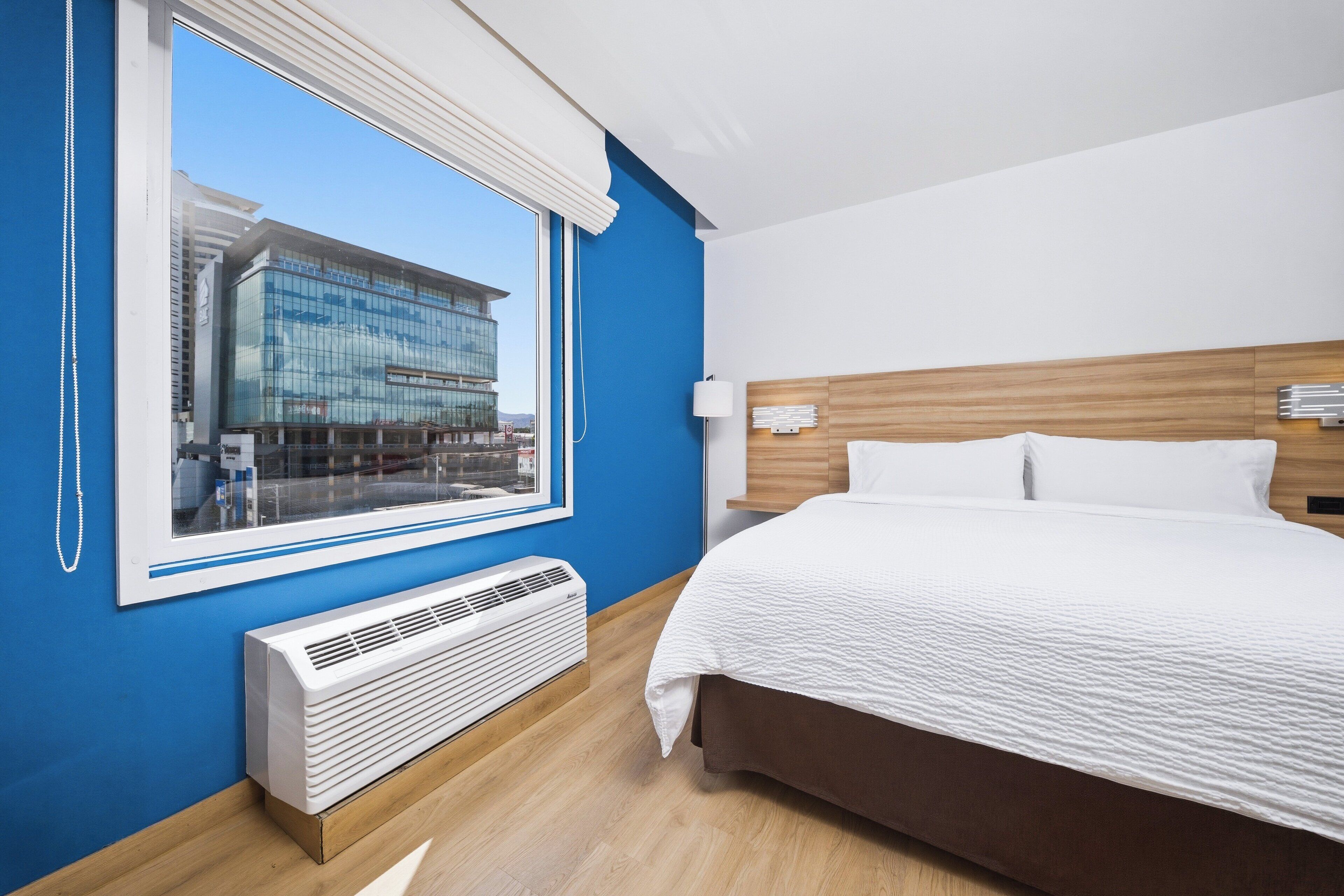 Quarto de hotel com uma janela grande, uma cama, mesa de cabeceira, luminária e aquecedor fixado na parede.