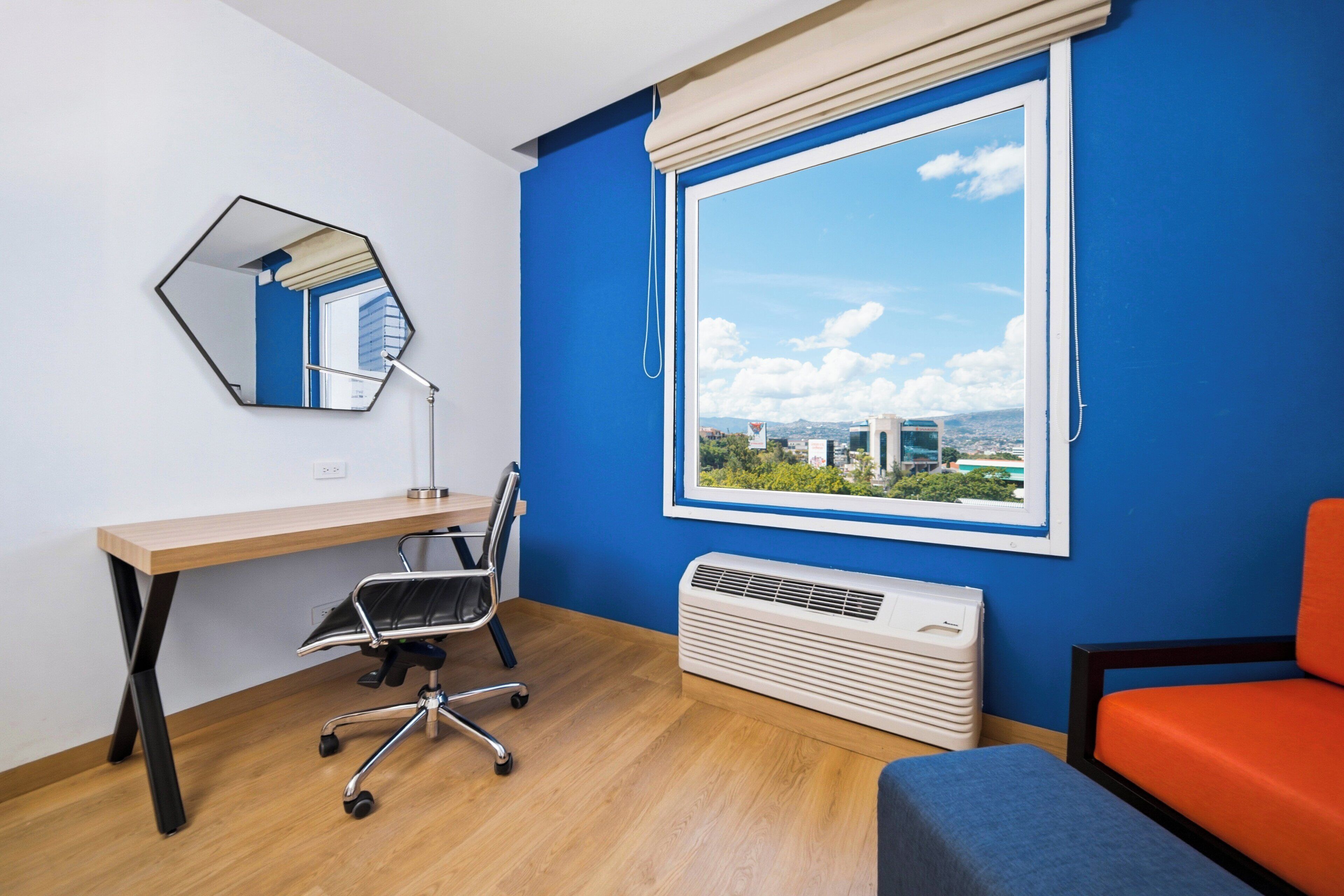 Una habitación con escritorio de madera, una silla, un ventanal con vistas, un espejo y un calentador.