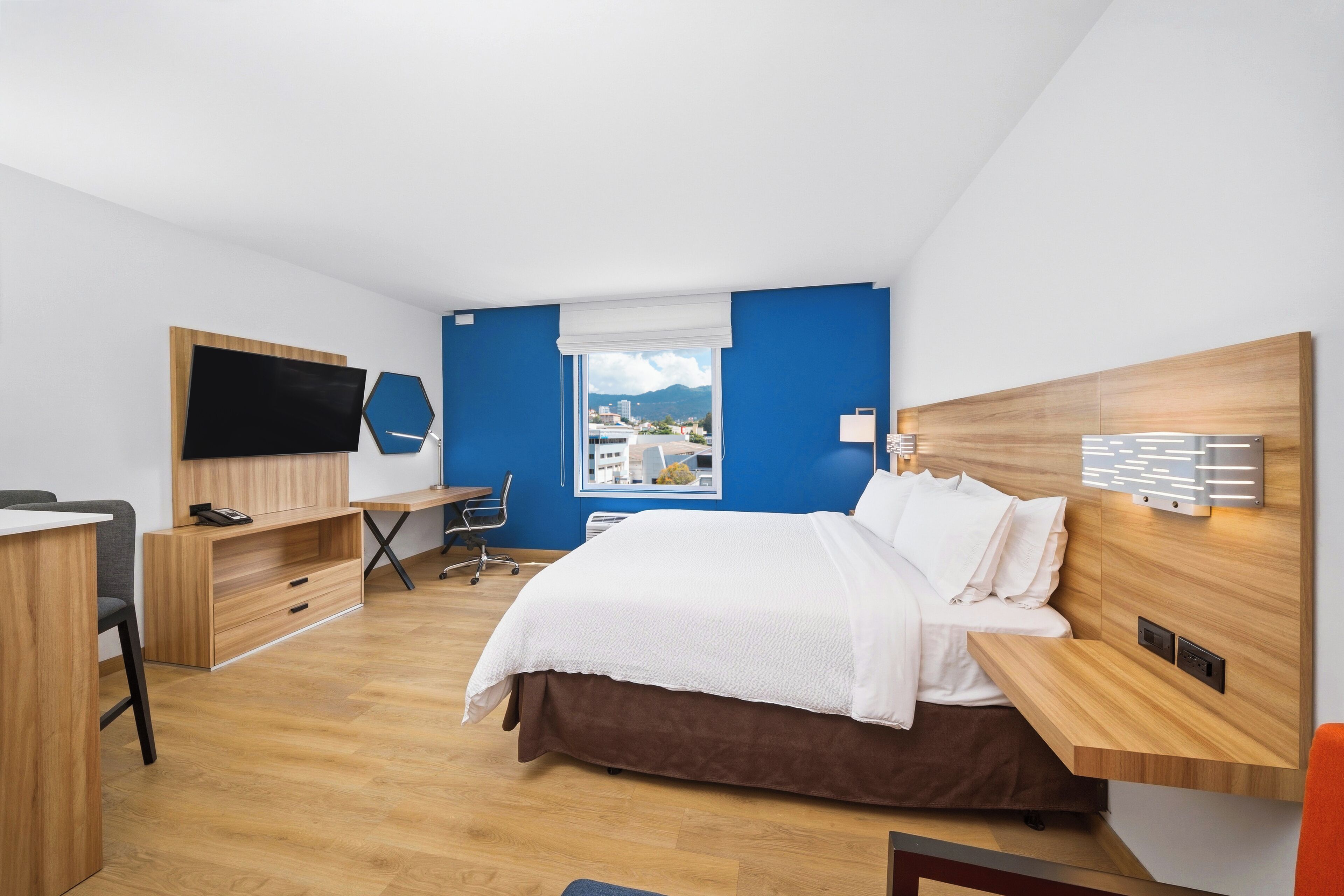 A modern hotel room with a bed, desk, TV, and a window with a city view.