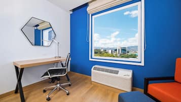 A room with a wooden desk, a chair, a large window with a view, a mirror, and a heater.
