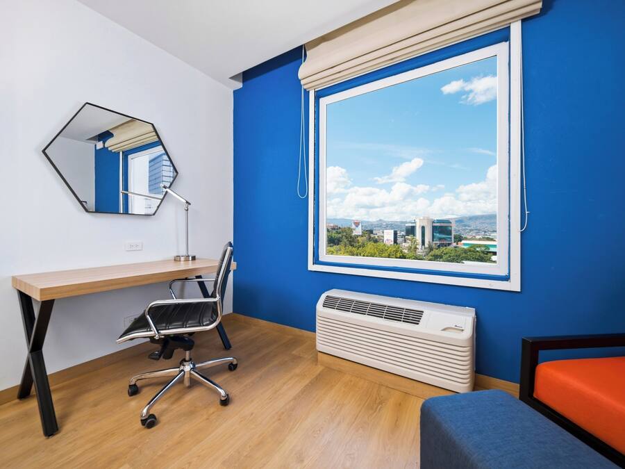 A room with a wooden desk, a chair, a large window with a view, a mirror, and a heater.