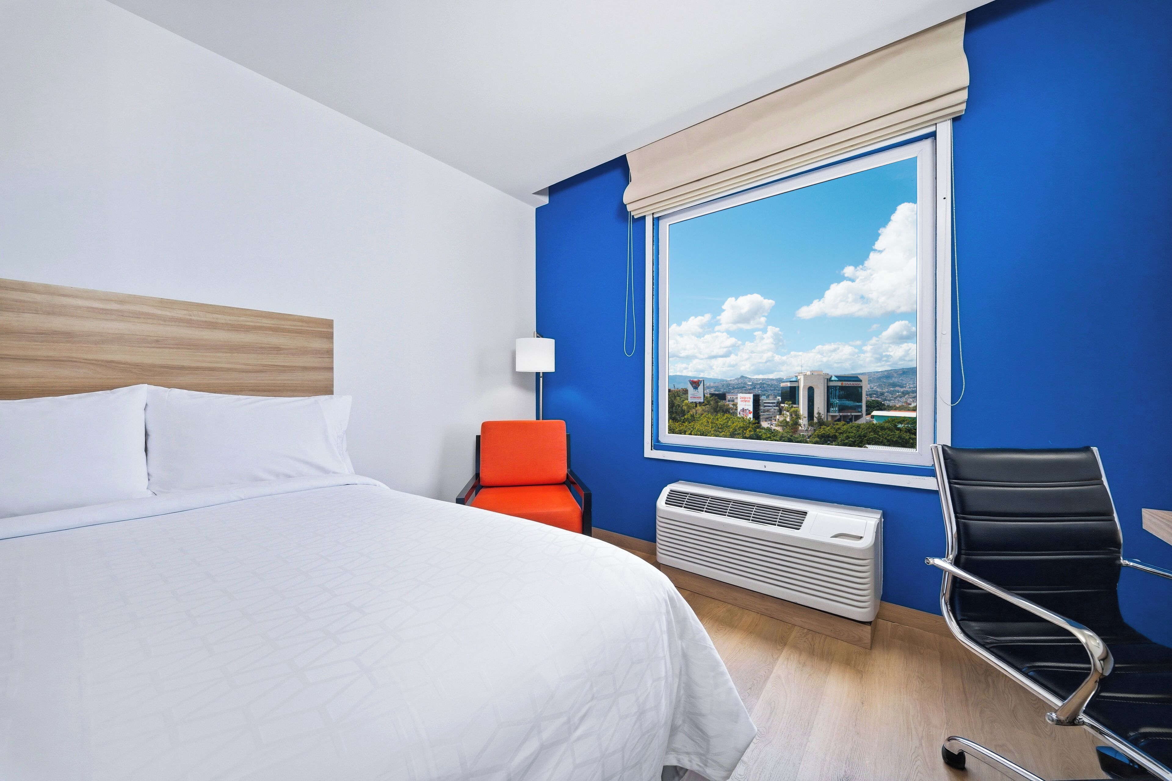 Una habitación de hotel con cama, una silla, un escritorio, un ventanal con vista a la ciudad y un radiador.