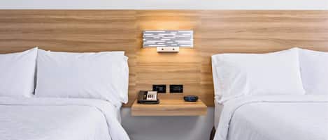 A hotel room with two beds, a wooden headboard, a bedside table with a phone and a digital clock, and a wall-mounted light fixture.