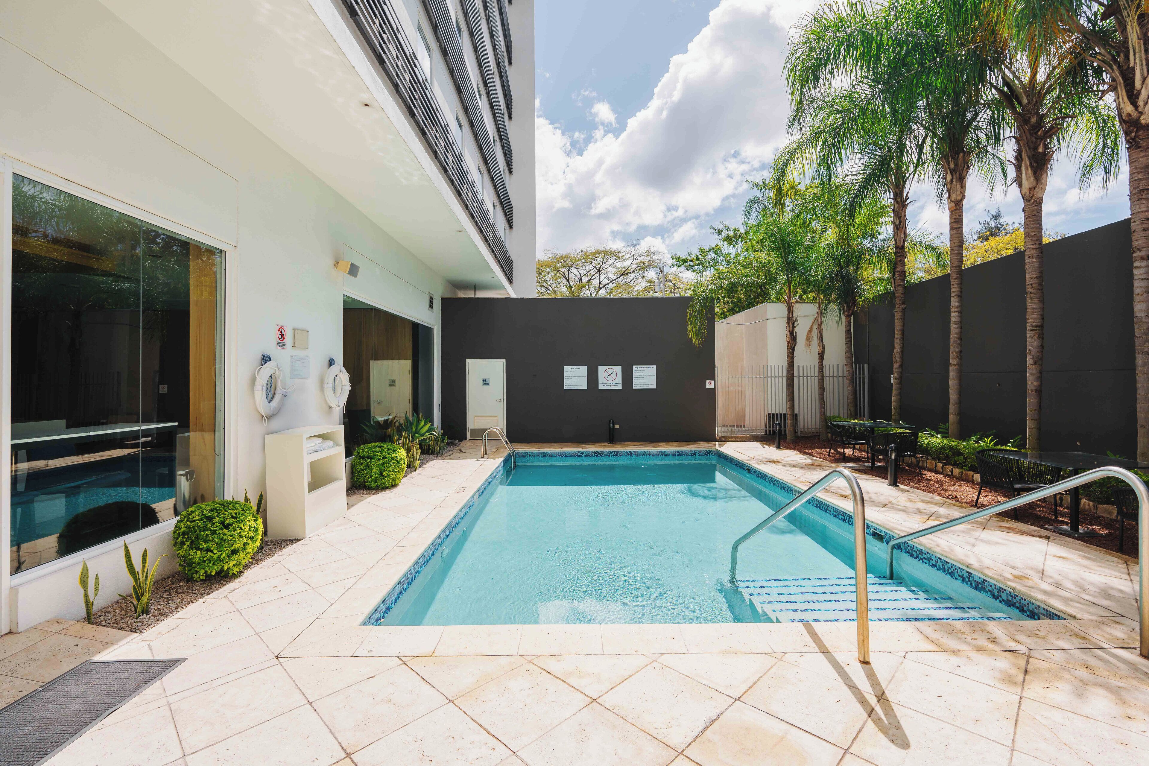 A swimming pool with a tiled deck, a covered area with seating, and a building with a dark facade.