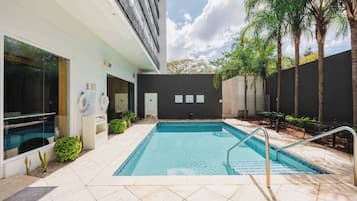 A swimming pool with a tiled deck, a covered area with seating, and a building with a dark facade.