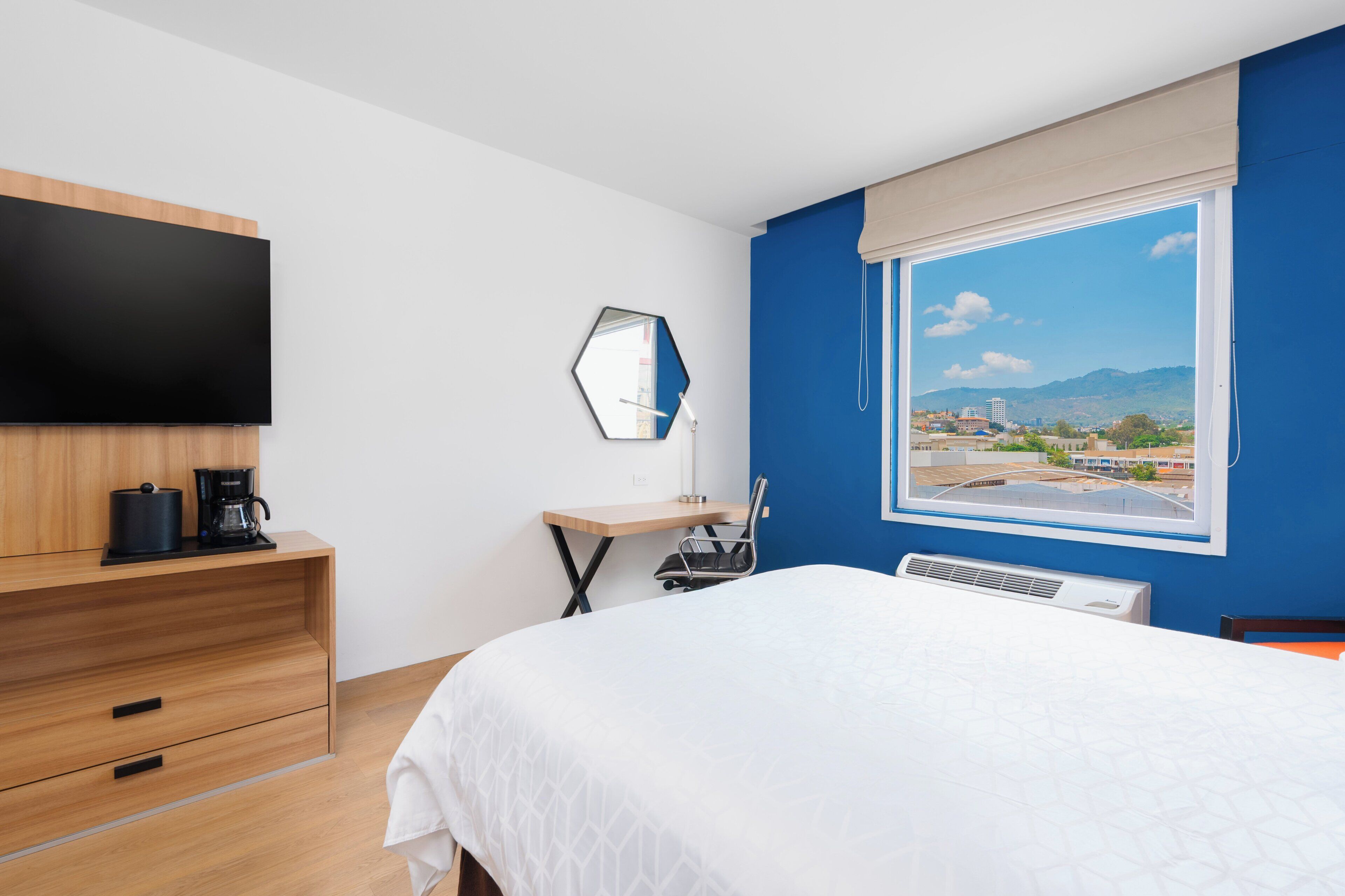 Habitación de hotel con cama, escritorio, televisor de pantalla plana, cafetera y ventana grande con vistas a la ciudad.