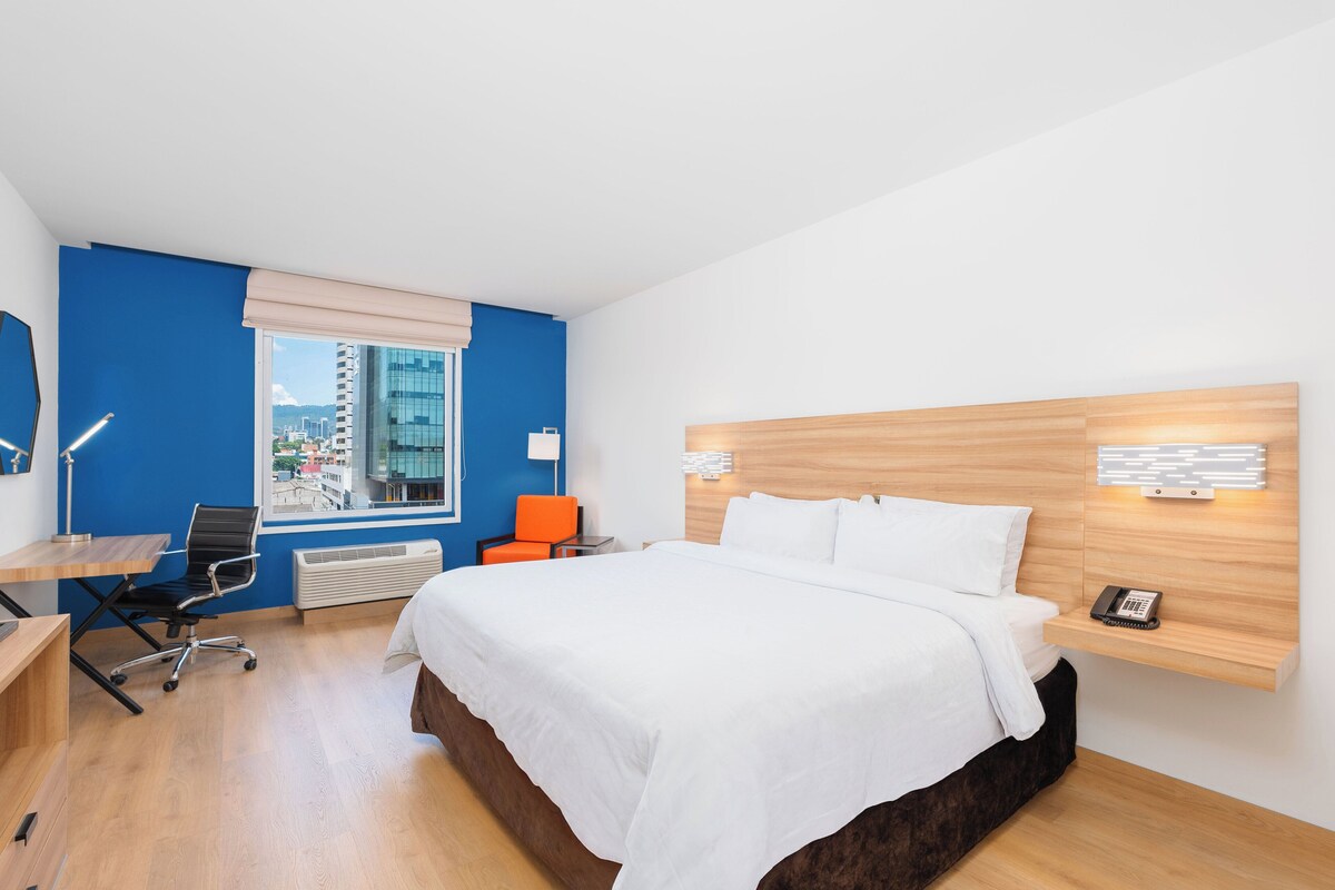 A modern hotel room with a large bed, a desk with a chair, a window with a city view, and a small table with a lamp.