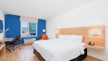Ein modernes Hotelzimmer mit einem großen Bett, einem Schreibtisch mit Stuhl, einem Fenster mit Stadtblick und einem kleinen Tisch mit Lampe.