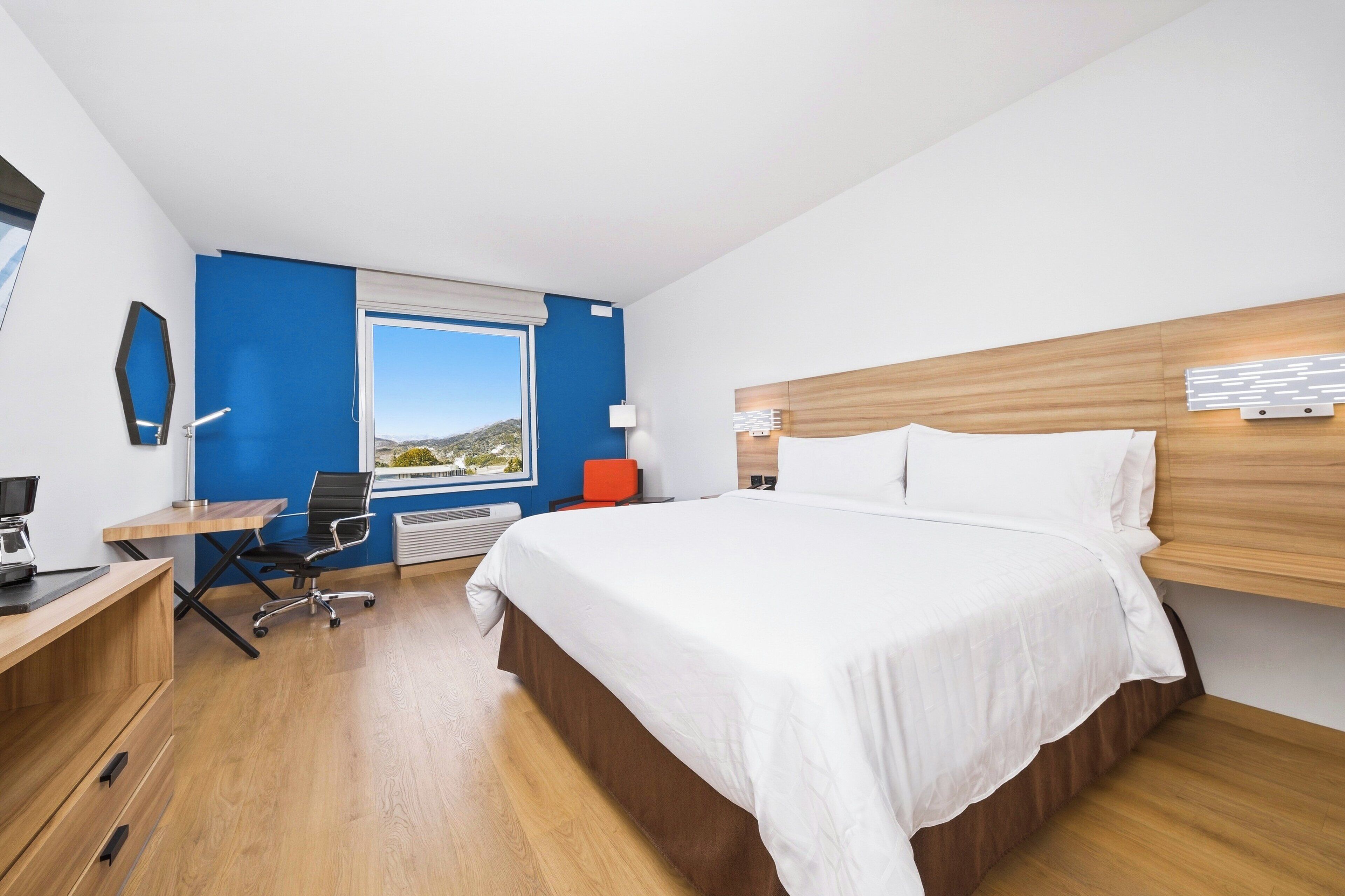 A modern hotel room with a wooden desk, a comfortable chair, a large bed, and a view of the outdoors.