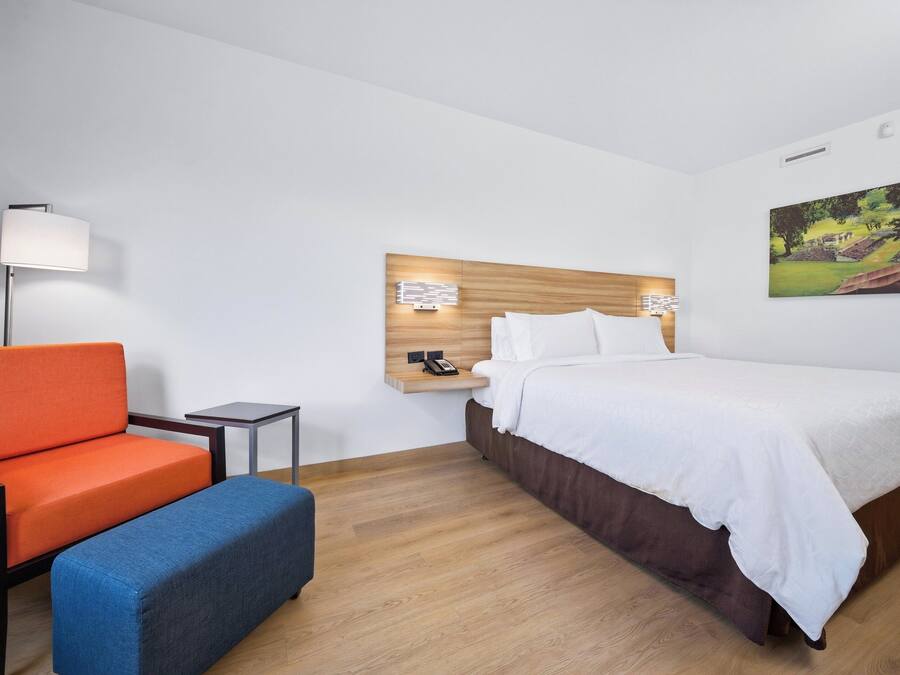 A hotel room with a bed, a nightstand, a lamp, a chair, and a bench.