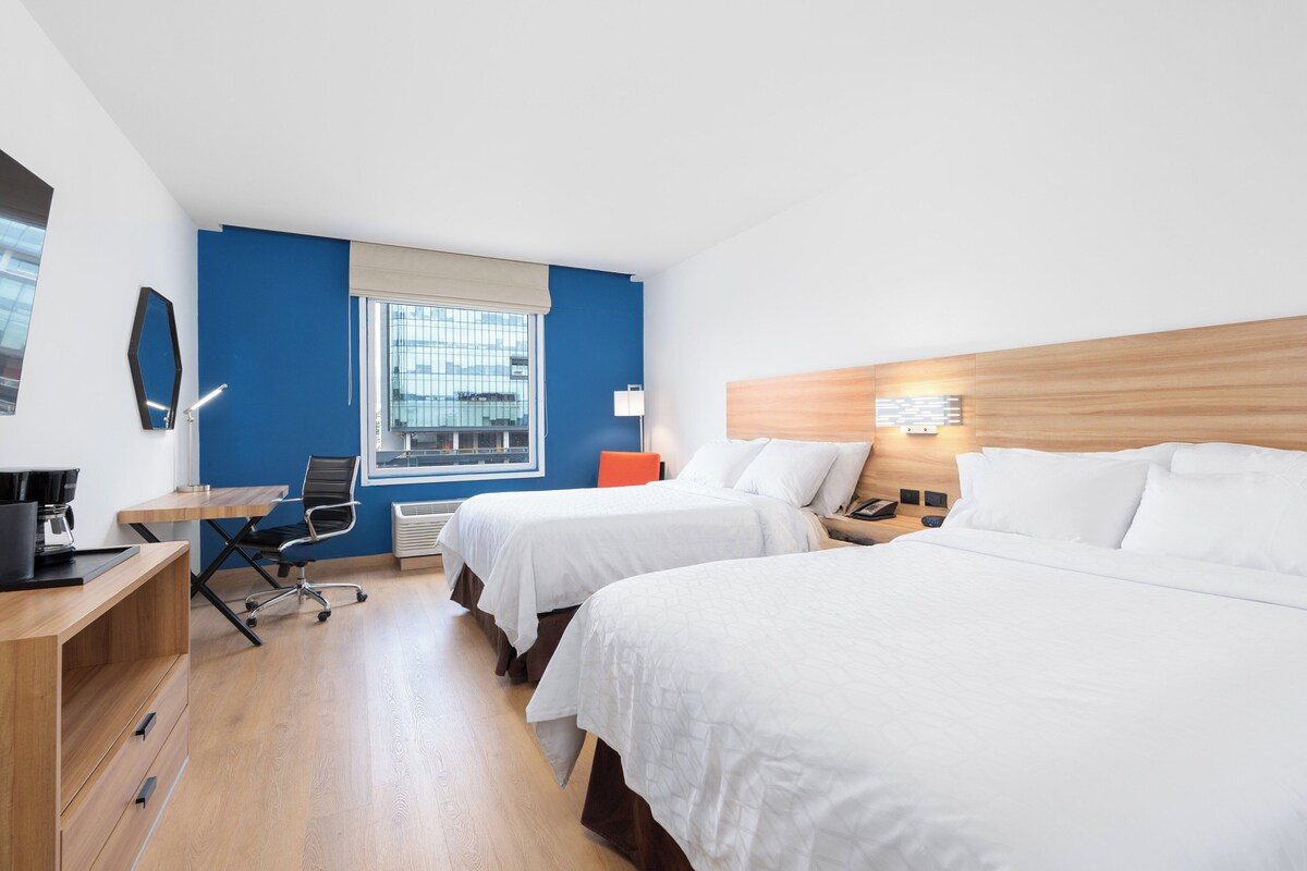 A hotel room with two beds, a desk with a chair, a lamp, and a window with blinds.