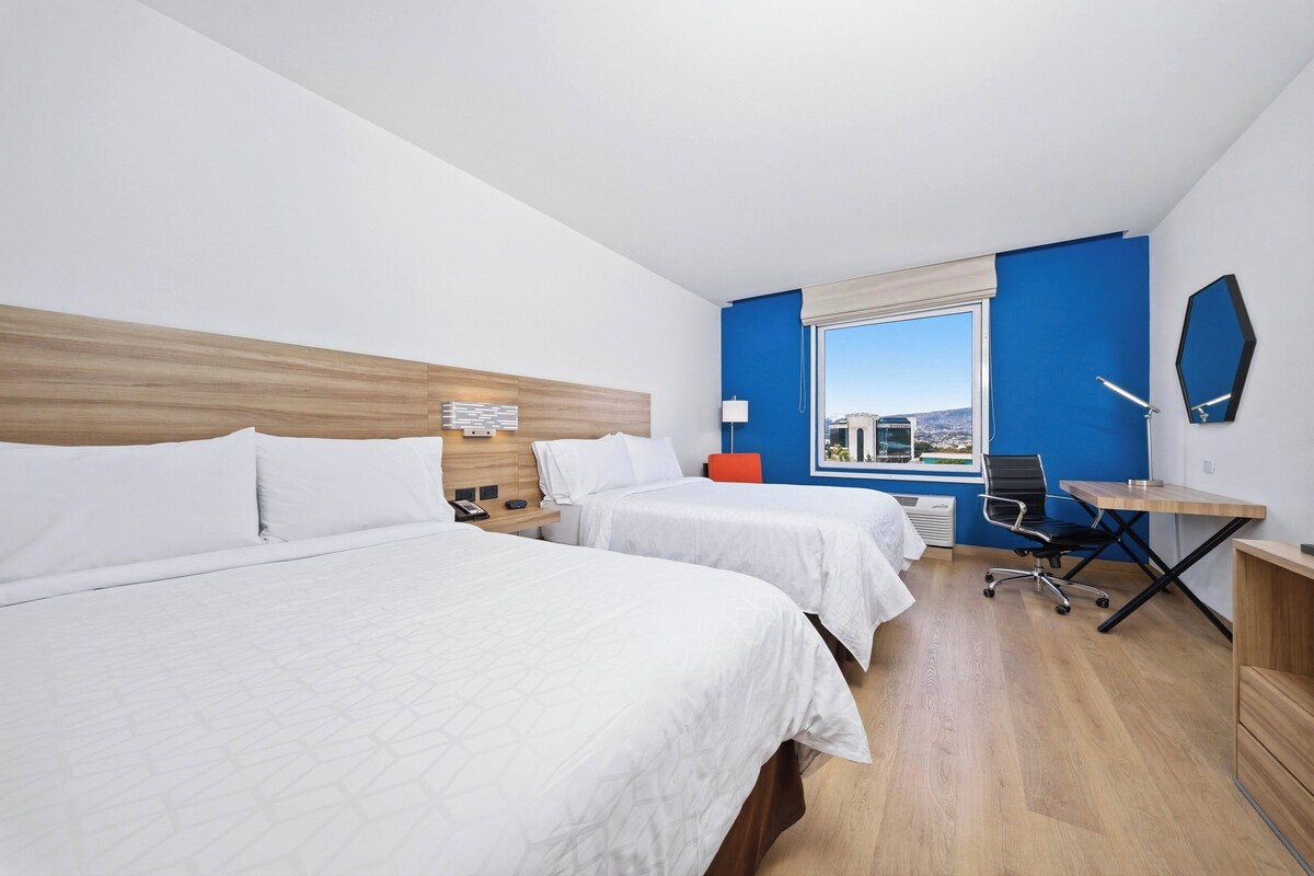 A hotel room with two beds, a desk, a chair, a window with a view, and a mirror on the wall.