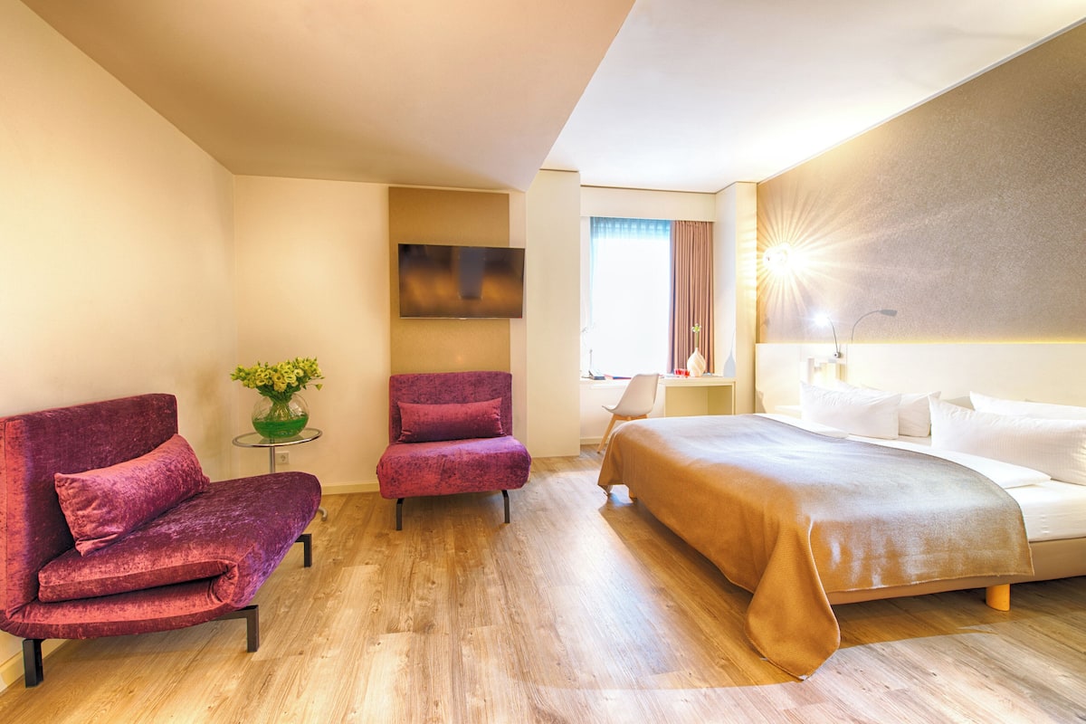 A hotel room with a bed, two armchairs, a small table with a vase, and a TV on the wall.