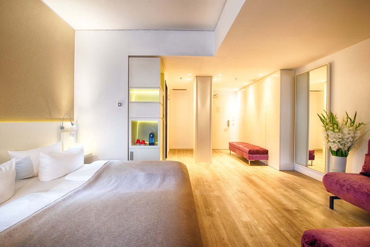 A modern hotel room with a large bed, a red bench, a mirror, and a plant.
